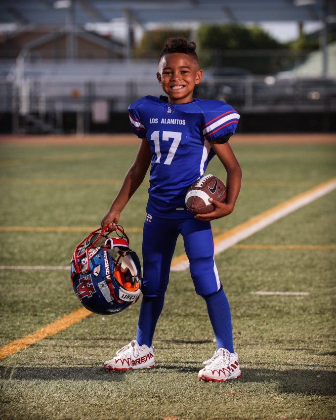 Young football player #17 in Los Alamitos blue uniform holding ball and helmet on field